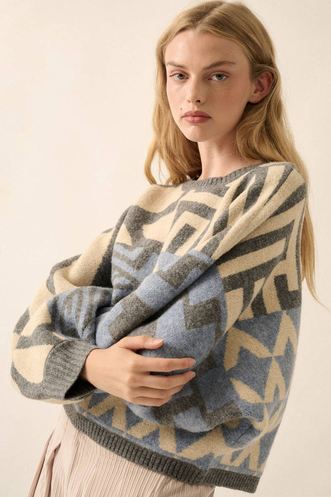 Model wearing a patterned knit sweater with bold geometric designs in neutral and contrasting tones, styled with casual bottoms.