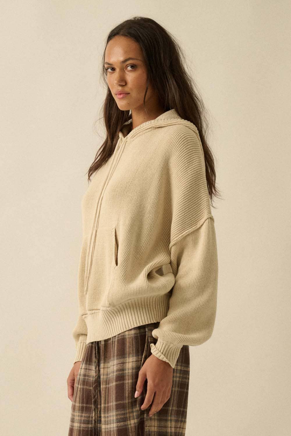 Woman wearing a beige hoodie and plaid pants against a plain background