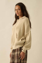 Woman wearing a beige hoodie and plaid pants against a plain background