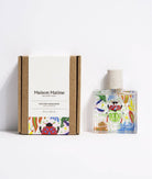 Maison Matine Nature Insolente perfume bottle with colorful leaf and beetle illustrations, placed on a vibrant yellow fabric and surrounded by green foliage.