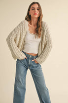 Woman wearing a loose cable knit cardigan with wide sleeves, paired with a white ribbed tank and blue denim jeans.