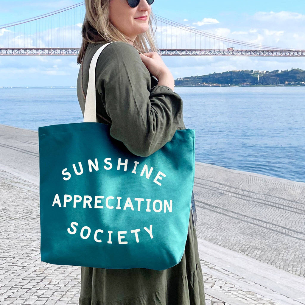Teal tote bag with “Sunshine Appreciation Society” in white text, held by a person in a green dress at the beach