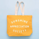 Yellow canvas tote bag with “Sunshine Appreciation Society” printed in white, held up by a hand in front of a sage green wall