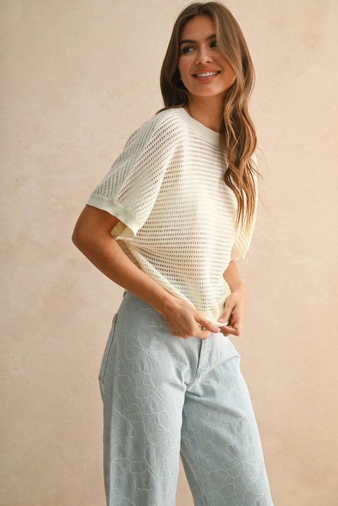 Model wearing a cream open-knit short sleeve sweater with open back detail, paired with embroidered light-wash denim pants.