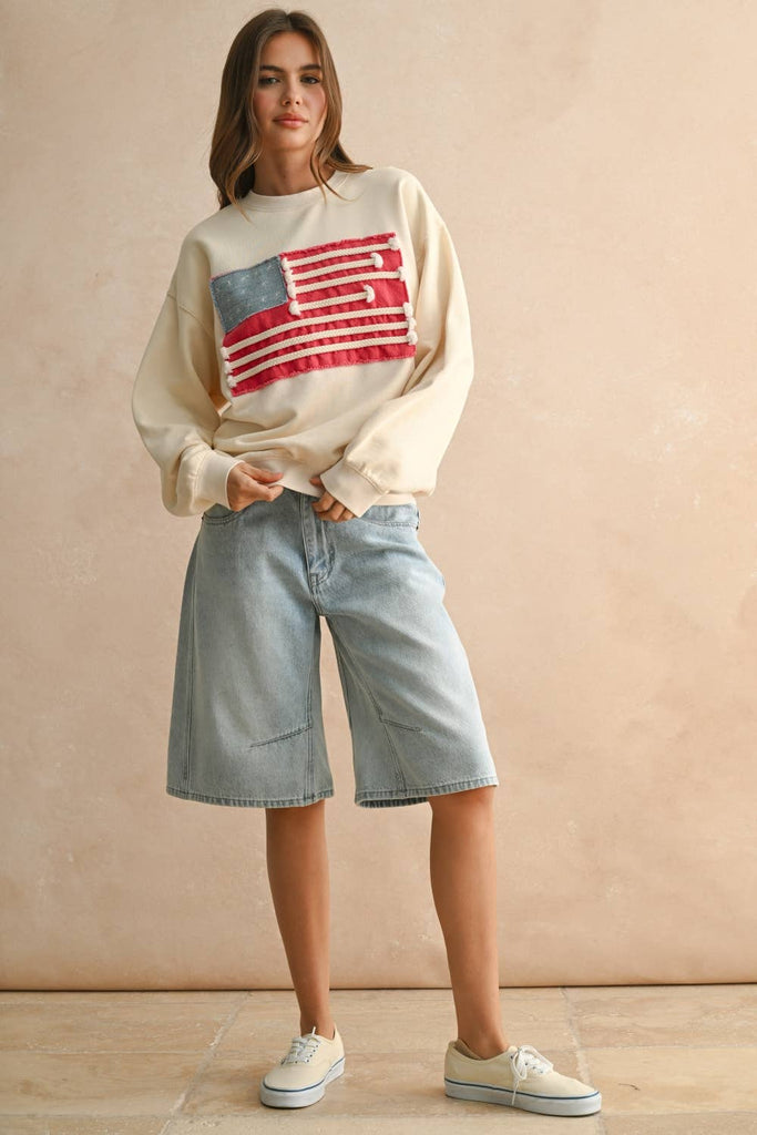 Woman wearing a cream sweater with a textured rope-stripe American flag design, paired with light wash jeans.