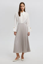 Model wearing a high-waisted pleated maxi skirt styled with a white blouse and pointed heels.