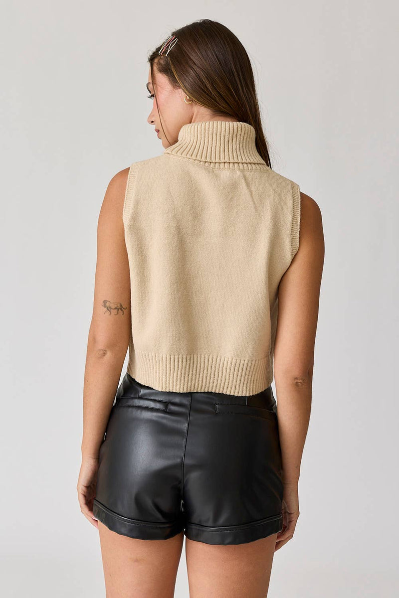 Woman wearing a beige sleeveless turtle neck knit cropped top with black leather shorts.