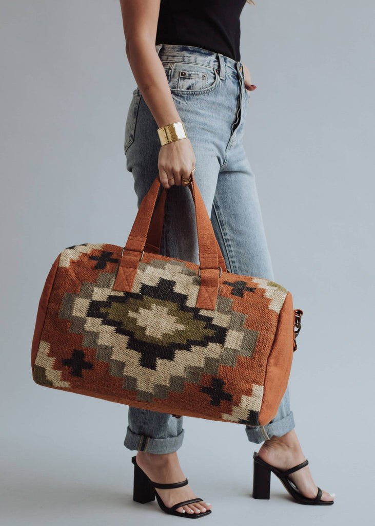 Earth-tone woven weekender bag with geometric pattern in black, green, and cream, held by woman in jeans and black top