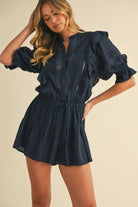 Woman wearing a navy cotton romper with puff sleeves, ruffle and lace details, and a drawstring waist.