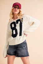 Woman wearing a cream knit sweater with a bold black "87" graphic and dark denim shorts.