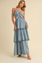 Woman wearing a denim maxi dress with a large front bow, side cutouts, spaghetti straps, and tiered skirt.