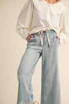 Woman wearing wide-leg light denim pants with a tie-front waist, paired with a white blouse and ballet flats.