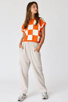 Woman wearing an orange and white loose-fit checker knit top with beige pants.