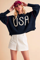 A woman wearing a navy sweater with the text 'USA' in white letters, paired with white shorts and a red cap.
