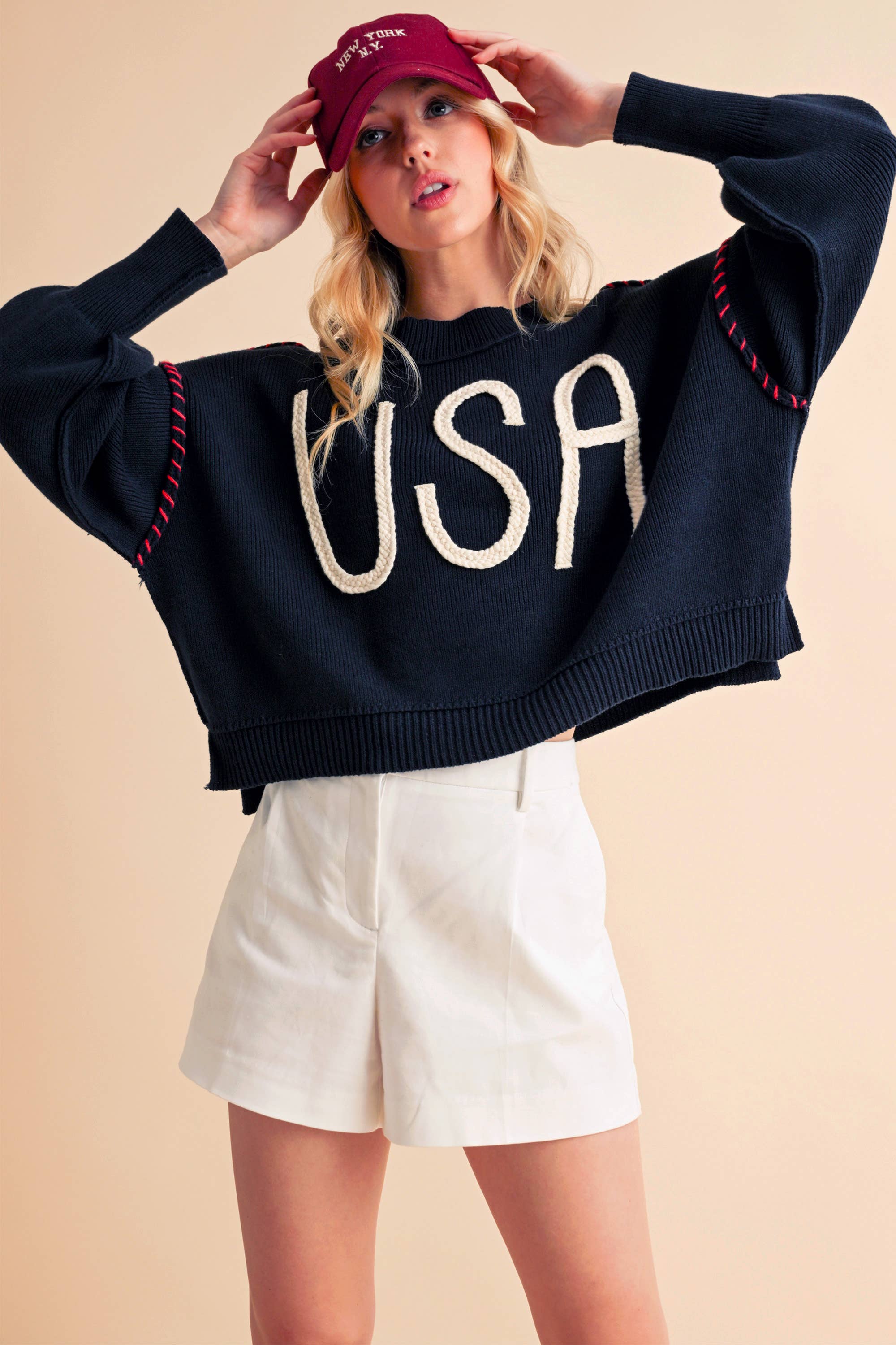 A woman wearing a navy sweater with the text 'USA' in white letters, paired with white shorts and a red cap.