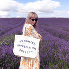 Cream canvas tote bag with “Sunshine Appreciation Society” printed in navy, filled with flip-flops, sunglasses, and travel essentials on a pink background