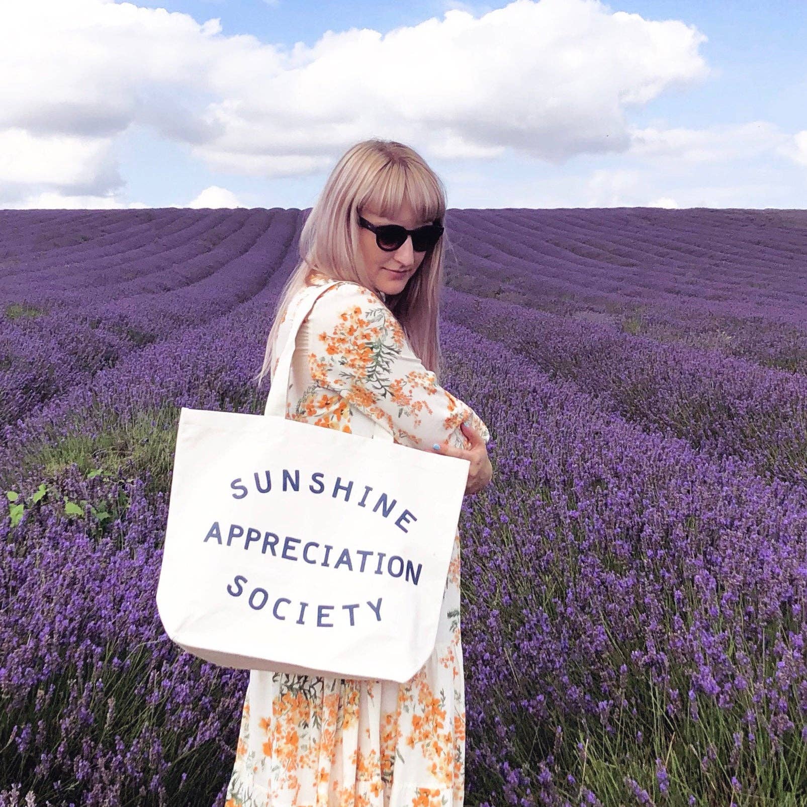 Cream canvas tote bag with “Sunshine Appreciation Society” printed in navy, filled with flip-flops, sunglasses, and travel essentials on a pink background