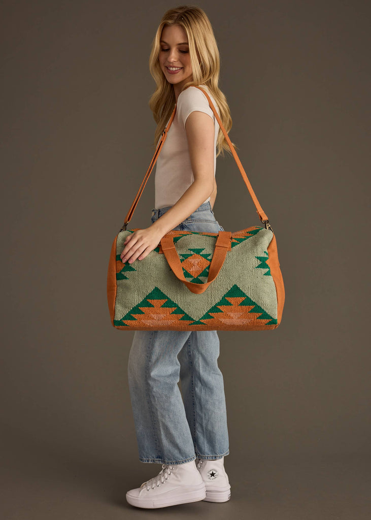 Sage green and rust weekender bag with geometric tribal pattern in green and orange, held by woman in jeans and white tank