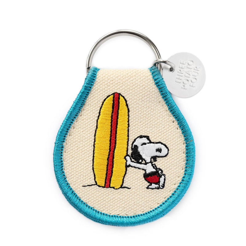 Peanuts® - Snoopy Surf Keychain with an embroidered design of Snoopy leaning on a yellow and red surfboard, framed with blue trim, and a metal “Three Potato Four” tag.