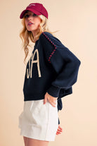 A woman wearing a navy sweater with the text 'USA' in white letters, paired with white shorts and a red cap.