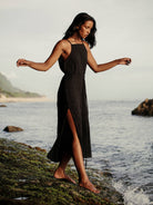 A woman standing on a rocky beach wearing a black maxi dress with spaghetti straps and a square neckline, with a slit on the left side of the dress.