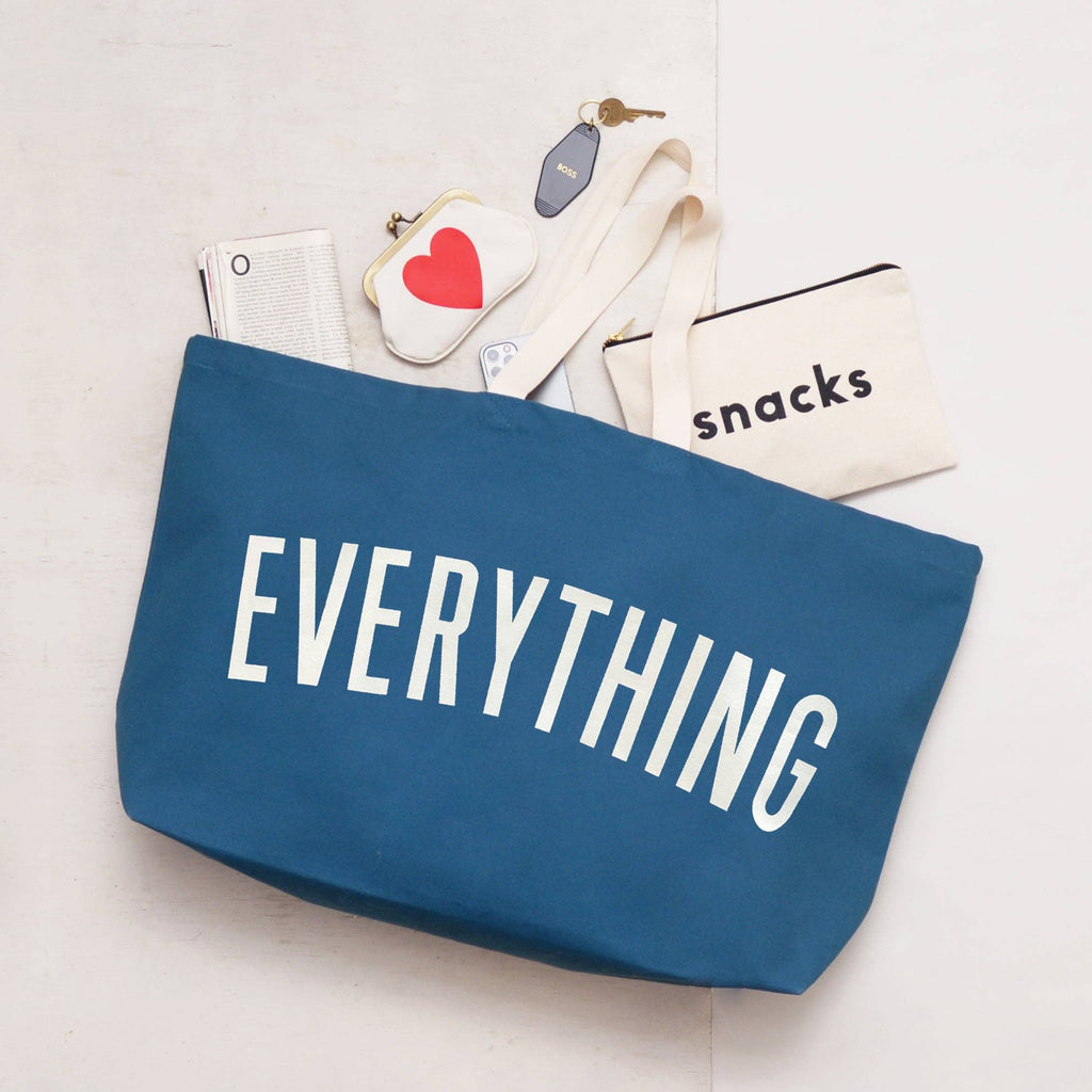 Oversized navy blue tote bag with cream handles and bold white “EVERYTHING” text, held by a hand against a light wood background