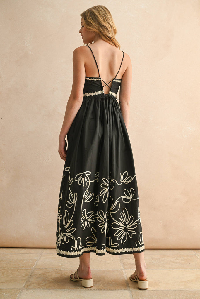 Woman wearing a black spaghetti-strap maxi dress with ivory floral embroidery and scalloped trim detail.