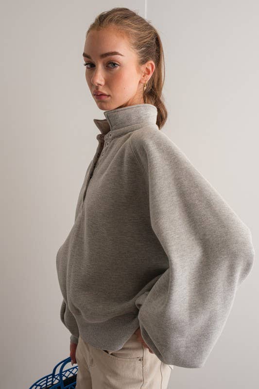 Woman wearing an oversized gray pullover sweatshirt with quarter snaps and voluminous sleeves, paired with beige pants.