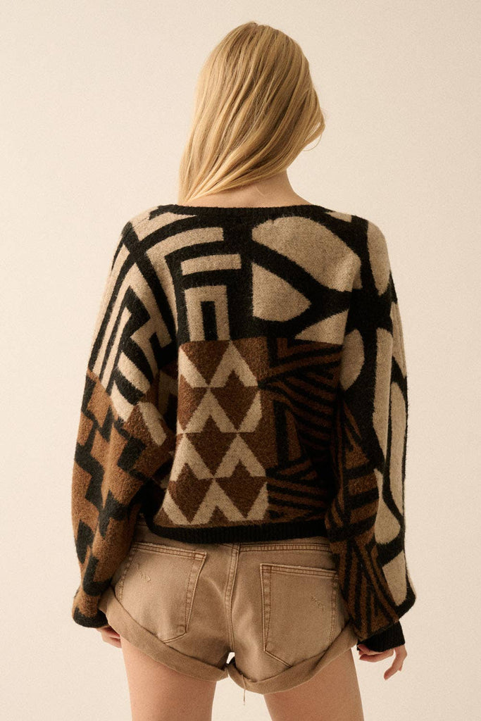 Model wearing a patterned knit sweater with bold geometric designs in neutral and contrasting tones, styled with casual bottoms.