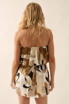 Woman wearing a strapless ruffle mini dress with smocked waist and bold abstract print in beige, black, and brown tones.