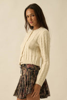 Woman wearing a cream textured knit cardigan with covered buttons, styled with a floral skirt.