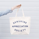 Cream canvas tote bag with “Sunshine Appreciation Society” printed in navy, filled with flip-flops, sunglasses, and travel essentials on a pink background
