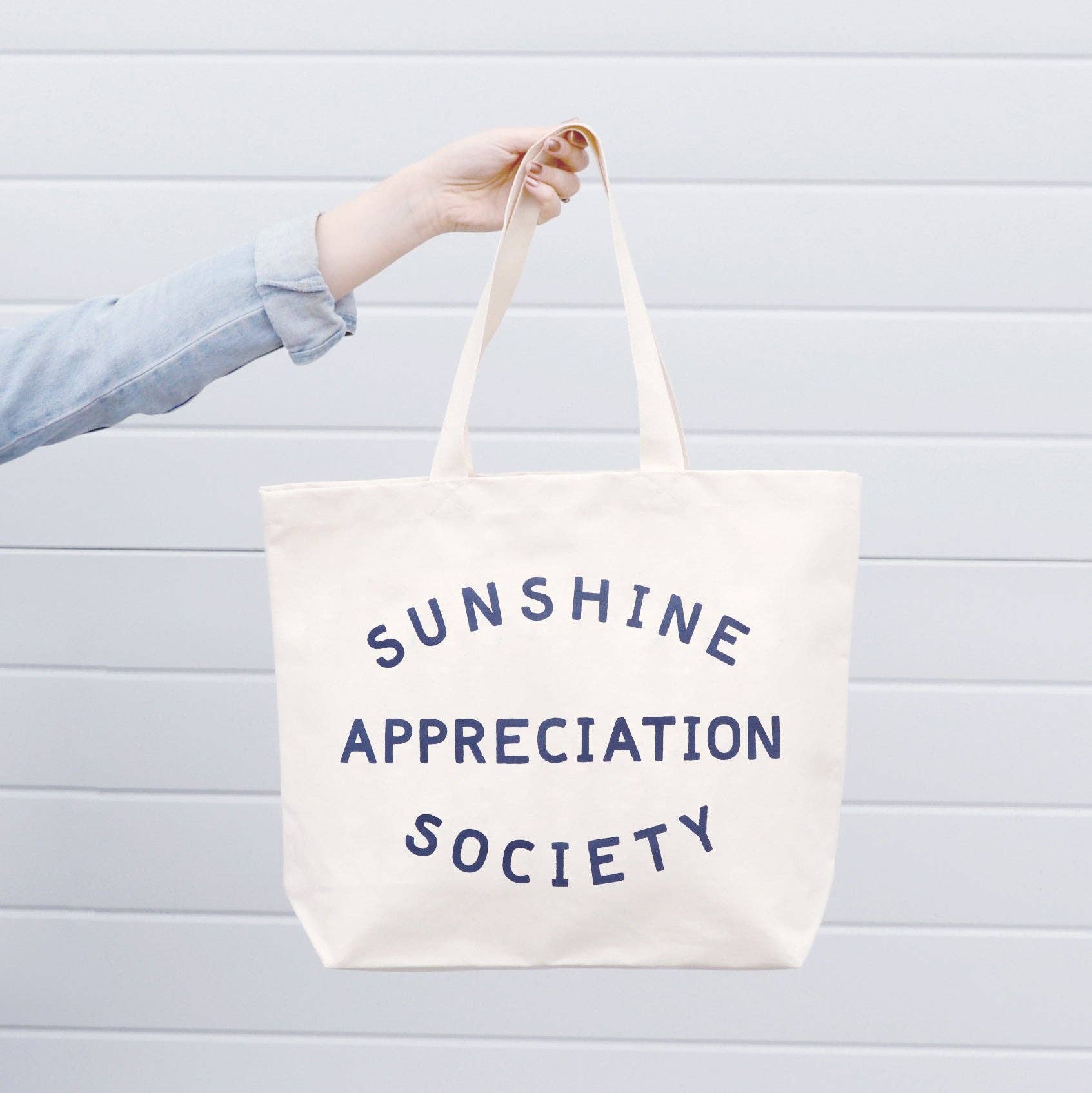 Cream canvas tote bag with “Sunshine Appreciation Society” printed in navy, filled with flip-flops, sunglasses, and travel essentials on a pink background