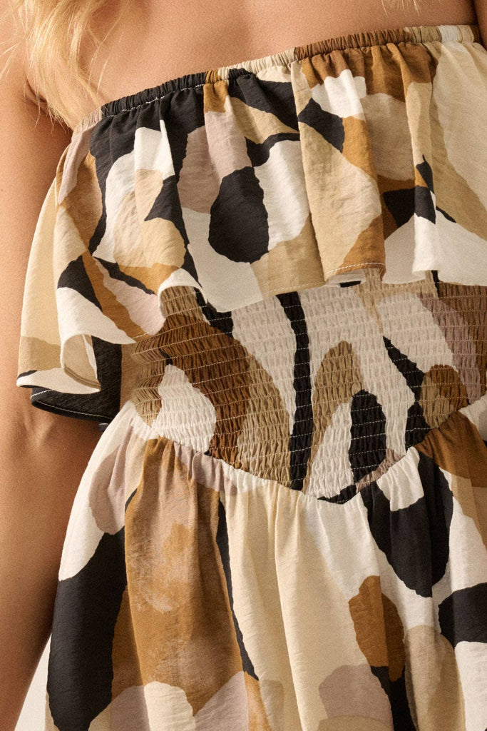 Woman wearing a strapless ruffle mini dress with smocked waist and bold abstract print in beige, black, and brown tones.