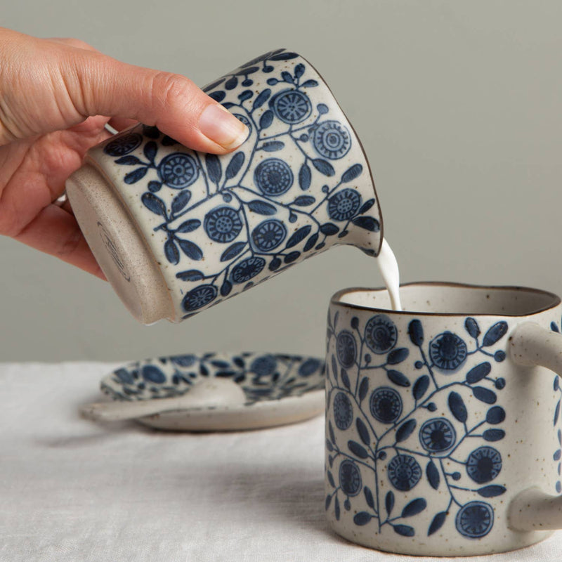Person pouring milk from a blue and white patterned ceramic pitcher into another similar mug on a light surface.