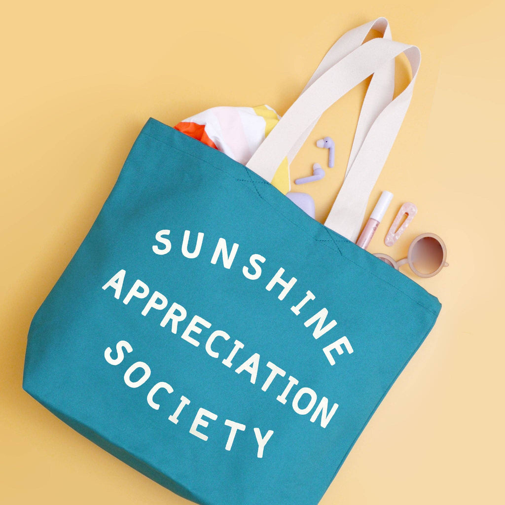Teal tote bag with “Sunshine Appreciation Society” in white text, held by a person in a green dress at the beach