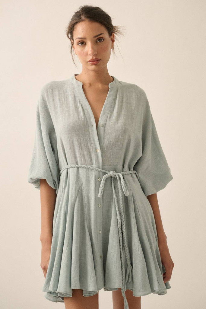 Woman wearing a sage green button-down mini dress with a braided waist tie and flowy hemline.