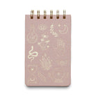 A pink cloth-covered notepad with a gold foil spiral binding and various gold foil accents on the cover, including symbols such as eyes, leaves, and celestial bodies.