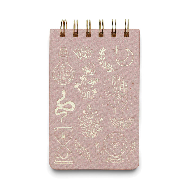 A pink cloth-covered notepad with a gold foil spiral binding and various gold foil accents on the cover, including symbols such as eyes, leaves, and celestial bodies.