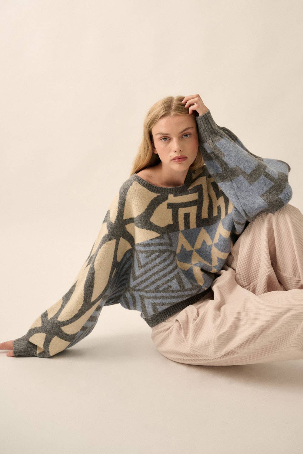 Model wearing a patterned knit sweater with bold geometric designs in neutral and contrasting tones, styled with casual bottoms.