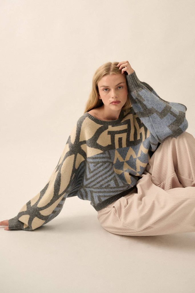 Model wearing a patterned knit sweater with bold geometric designs in neutral and contrasting tones, styled with casual bottoms.