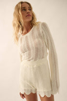 Woman wearing a beige knit romper with bell sleeves, scalloped hems, and open-weave detailing.
