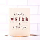Off-white card with black and pink text saying ‘You’re Weird & I Love You,’ presented with a brown envelope.