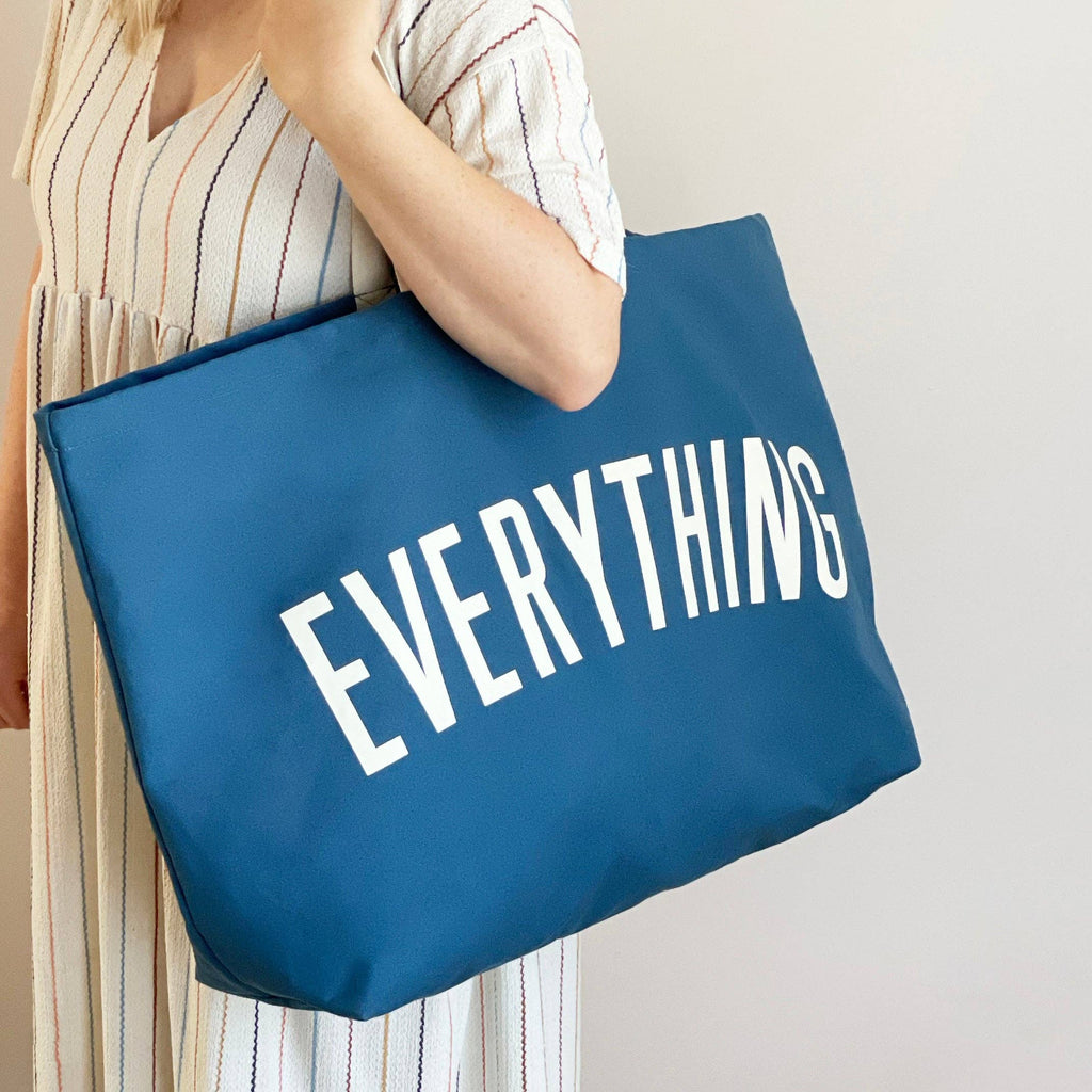 Oversized navy blue tote bag with cream handles and bold white “EVERYTHING” text, held by a hand against a light wood background