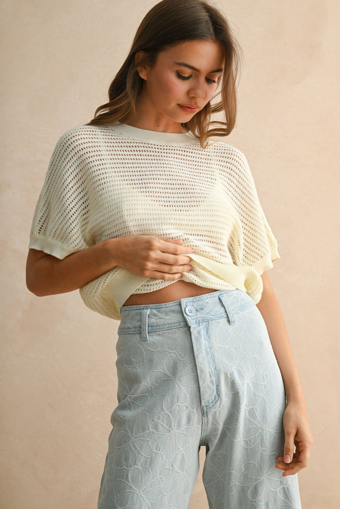 Model wearing a cream open-knit short sleeve sweater with open back detail, paired with embroidered light-wash denim pants.