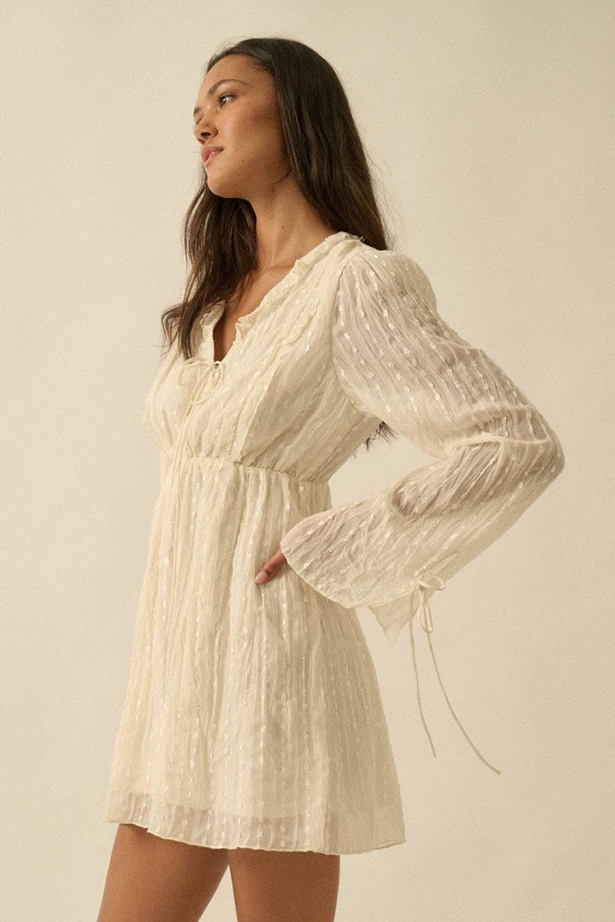 Woman wearing a cream mini dress with sheer long sleeves, button front, and tie details.