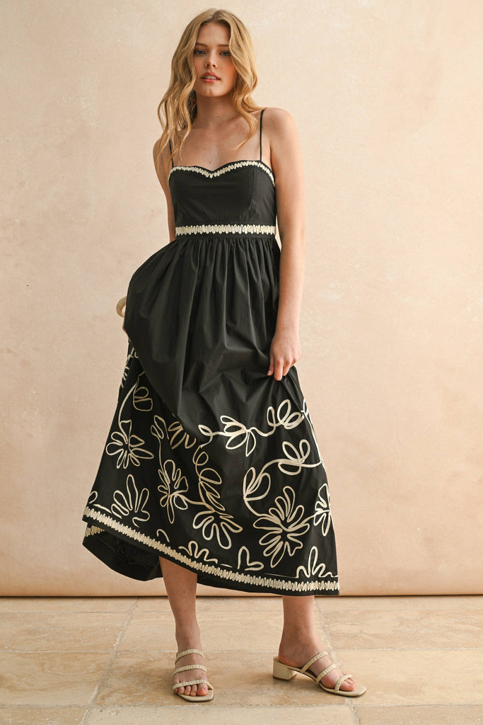 Woman wearing a black spaghetti-strap maxi dress with ivory floral embroidery and scalloped trim detail.