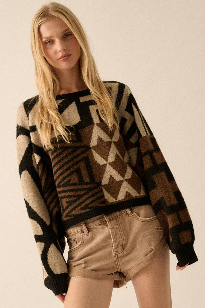 Model wearing a patterned knit sweater with bold geometric designs in neutral and contrasting tones, styled with casual bottoms.