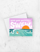 Surf-themed birthday card with large wave, surfers, and a sunset with the text “Hope your birthday’s swell”.