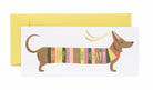 Happy Birthday greeting card with a dachshund wearing a colorful striped sweater, gold foil lettering, and a yellow envelope.